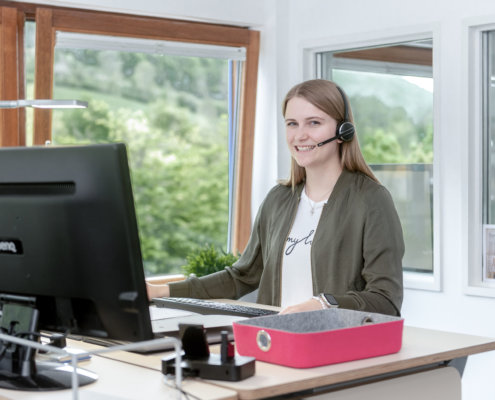 Junge Frau sitzt lächelnd mit einem Headset am Computer Bildschirm und arbeitet